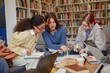 © Seventyfour - Three young adult girls sitting together studying and writing in notebooks, collaborating on academic project in library with laptops and coffee cups visible