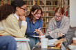 © Seventyfour - Group of multiethnic teenage girls collaborating on school project in library, discussing notes and studying together with books and stationery on table, focused on teamwork and learning