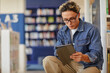 © Seventyfour - Young adult man sitting in library reading digital tablet, wearing glasses and focusing on screen, surrounded by bookshelves and blurred background