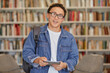 © Seventyfour - Portrait of Asian boy wearing glasses smiling and holding digital tablet in library setting, carrying backpack and looking directly at camera, bookshelves in background