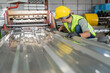 © tong2530 - A Worker in safety gear inspecting the quality of a corrugated metal sheet (roofing panel) as it is produced by a roll forming machine in a factory.