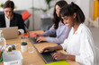 © Studio Marmellata - Three diverse women collaborate in a modern office, each focused on their laptops. The smiling Black woman in the foreground actively types, alongside her concentrating colleagues.