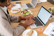 © Studio Marmellata - Two people work at a wooden table. One types on a laptop, another writes in a notebook. Healthy lunch items like salad and sandwiches are on the table, showing a productive session.