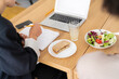 © Studio Marmellata - Two people enjoy a healthy lunch break at a modern wooden table. One person is actively writing in a notebook, with a laptop, sandwich, fresh salad, and a drink nearby.