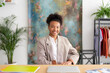 © Studio Marmellata - A smiling young Black woman sits at a desk in a bright office. She wears a plaid blazer, looking confidently at the camera. Her workspace includes a laptop and colorful folders.