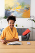 © Studio Marmellata - A smiling young Black woman sits at a desk in a modern home office. She has a laptop, notebook, and phone on the wooden surface, ready for work or study.
