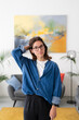 © Studio Marmellata - A young woman with glasses stands confidently in a modern living room, looking at the camera. She wears a denim shirt and has her hand behind her head.
