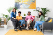 © Studio Marmellata - Four diverse young women are smiling and sitting comfortably in a modern living room. They are enjoying a casual gathering at home, looking at the camera.