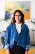 © Studio Marmellata - A young woman with long brown hair and glasses stands confidently indoors, looking directly at the camera. She wears a blue denim shirt over a white t-shirt, portraying a casual yet focused demeanor.