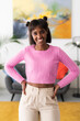 © Studio Marmellata - A smiling young Black woman with space buns stands confidently in a modern living room, hands on hips. She wears a pink cable-knit sweater and light pants, looking at the camera.