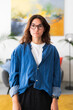 © Studio Marmellata - A young woman with brown hair and glasses stands indoors, looking directly at the camera. She wears a denim shirt, white t-shirt, and dark pants in a modern room with abstract art.