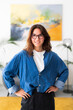 © Studio Marmellata - A cheerful young woman with dark hair and glasses smiles at the camera, standing confidently with hands on her hips. She is in a bright, modern room with an abstract painting.