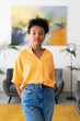 © Studio Marmellata - A confident young Black woman with curly hair stands in a modern indoor space, looking directly at the camera. She wears a vibrant yellow shirt and blue jeans.