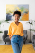 © Studio Marmellata - A smiling young Black woman with curly hair stands in a modern interior. She wears a yellow top and blue jeans, looking confidently at the camera.