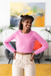 © Studio Marmellata - A smiling young Black woman stands confidently indoors with hands on her hips. She wears a pink sweater and light pants in a bright, modern living room.