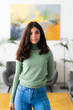 © Studio Marmellata - A young woman with long dark hair stands confidently in a modern living space. She wears a green turtleneck and blue jeans, looking directly at the camera.