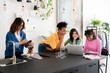 © Studio Marmellata - Four diverse young women gather in a modern space. One woman makes pour-over coffee while three friends smile and interact with a laptop, sharing a moment.
