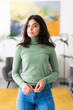 © Studio Marmellata - A young woman with dark hair stands indoors, looking at the camera. She wears a green turtleneck and blue jeans, with her hands clasped. The background features a modern living space.