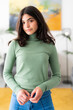 © Studio Marmellata - A young woman with dark hair stands indoors, looking at the camera. She wears a green turtleneck and blue jeans, hands clasped. A bright, modern living space is in the background.