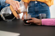 © Studio Marmellata - A person's hand, wearing a ring, pours coffee from a glass carafe into a small glass. Another person's hand, with painted nails, holds the glass, ready to receive the beverage.
