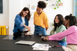 © Studio Marmellata - Four diverse young women gather around a table. One woman pours coffee into glasses while the others watch and smile, enjoying a casual social moment together.