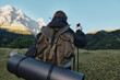 © SHOTPRIME STUDIO - A lone adventurer wearing a sturdy backpack marches across a wide meadow toward looming mountains under a clear sky, preparing for a hike and outdoor exploration.