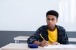 © Eve - Teen student sitting at desk in classroom, focused on writing test or assignment. Back to school, education and exam preparation concept. Horizontal indoors shot.