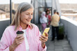 © Maria Vitkovska - Happy mature woman holding mobile phone, using mobile app cup with coffee outdoors on the street