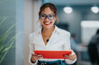 © qunica.com - Cheerful Filipino businesswoman stands near a glass wall, holding a bright red tablet. She wears glasses and a white blouse, exuding confidence and friendly collaboration in a corporate environment.