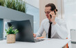 © qunica.com - A professional man sits at a clean, bright desk in a modern office, talking on a mobile phone while jotting notes beside a laptop. The scene conveys focus and productivity.