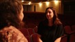 © PirisaTK - Two women engaged in conversation in an empty theater, surrounded by rows of red seats and soft lighting, creating an intimate atmosphere.
