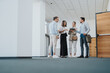 © qunica.com - A diverse group of colleagues stands in a bright office corridor, sharing a tablet and notes as they plan a project. The scene conveys teamwork, communication, and modern business culture.