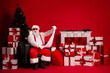 © deagreez - Santa reads a long list among gifts beside a Christmas tree against a bold red backdrop with wrapped presents and festive decor