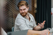 © qunica.com - A bearded man in a striped shirt participates in a business meeting, using expressive hand gestures in a contemporary office with colleagues and laptops nearby.