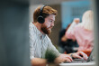 © qunica.com - A bearded man wearing large over-ear headphones concentrates on his laptop at a modern desk. The blurred background shows colleagues, conveying a busy, tech-focused office environment.