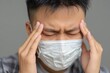 © LimeSky - Young Asian man in a mask experiences intense headache rubbing his temples with eyes closed looking distressed