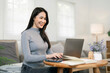 © NAMPIX - A beautiful and cheerful young Asian woman smiling while working or studying online with a laptop and notebook at a coffee table in her modern home.