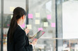 © PRIME STOCK LAB - Young professional woman planning project with sticky notes on glass wall while holding tablet