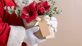 Santa Claus close-up hand in white glove holding bouquet of red amaryllis  eucalyptus in kraft paper box with blank tag, beige background, Christmas floral gift concept, copy  space flower delivery