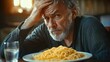 © tiger - Concerned middle-aged man with gray beard sitting at table with plate of spaghetti and glass of water, expressing frustration or worry indoors with warm lighting