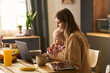 © pressmaster - Caucasian young adult woman working on laptop while holding toddler girl on lap in kitchen, both looking at screen, breakfast items and tablet on table, natural morning light streaming in