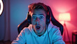 © Pete - Young man wearing headphones sits in chair, surprised expression on face. Gamer reacts to online game. Male streamer with open mouth looks at camera. Neon lighting in dark room, lamp in background.