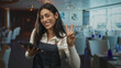 © Krakenimages.com - Woman holding tablet and flashing peace sign in restaurant dining room while wearing apron and smiling, tapping screen; service hospitality joy.