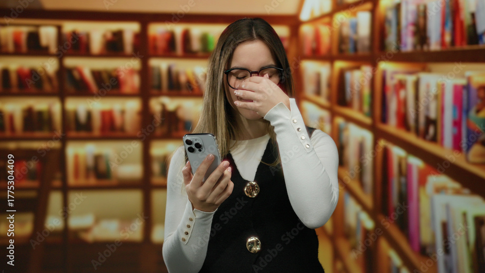 Woman holding smartphone in library with blonde hair and eyeglasses, showcasing a reflective mood under warm lighting among rows of bookshelves indoors.