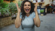 © Krakenimages.com - Woman in light blue shirt covering eyes with hands and wide open mouth on sunny street lined with green plants; surprise excitement.