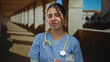© Krakenimages.com - Young female doctor in blue scrubs with stethoscope shrugs shoulders in old town buildings; disgust.