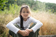 © PeterPike - A smiling girl in glasses sitting in the forest, looking down thoughtfully with a gentle expression. Blurred golden grass and sky in the background, soft natural light, copy space.