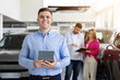 © Anastasiya - Happy car salesman standing with tablet in dealership, couple in background checking documents. Confident and successful sales representative in auto showroom