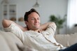 © ryker - Relaxed man reclining on sofa in bright modern living room with soft natural light and neutral decor, content expression and casual shirt creating calm comfortable home atmosphere