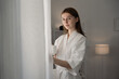 © Thitiporn - Young woman in white bathrobe standing by window with cup of coffee, enjoying peaceful morning light at home. Perfect concept of relaxation, slow living. woman, bathrobe, coffee, morning. cozy minimal